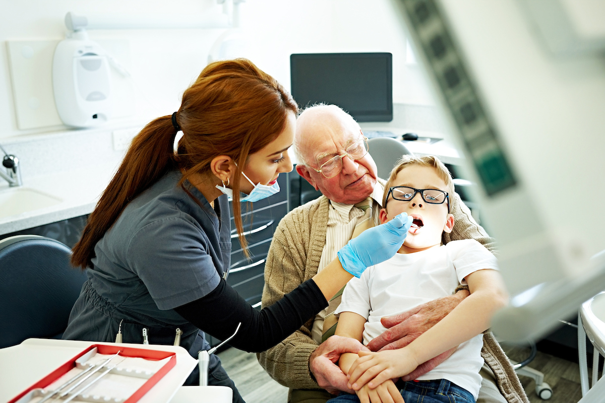 Dental assistant performing a check up on a child patient while his grandfather holds onto him