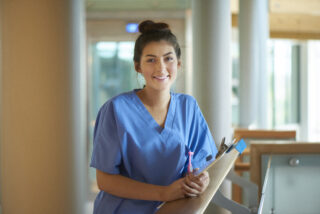 a young female nurse stands proudly on the hospital corridor . She is wearing scrubs and holding a medical records clipboard .