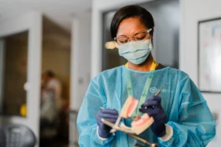 A dental assisting student in protective gear practices using dental instruments on a model of teeth.