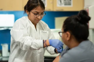A healthcare student in a lab coat and gloves practices a clinical procedure on a classmate’s arm in a training lab.