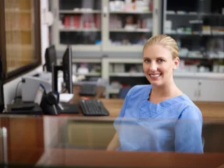 A smiling pharmacy professional in blue scrubs sits at a counter, with a computer and medication shelves behind her.